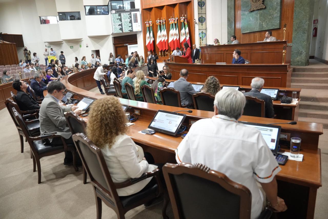 Diputados del estado de Sinaloa dentro del congreso en plena sesión, se ve a los diputados sentados en sus respectivos lugares y la bandera de México en la parte central del recinto