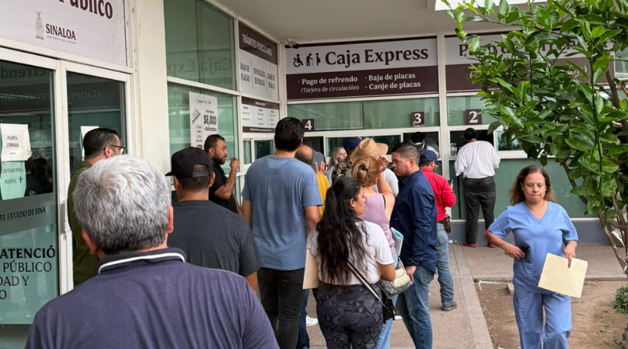 Un grupo de personas haciendo fila en las oficinas de recaudación del estado de Sinaloa, se ve al fondo a alguien realizando tramites y a una mujer caminando por la derecha saliendo del lugar