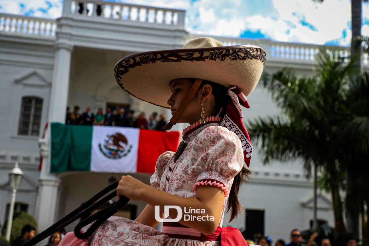 Niña disfrazada de adelita arriba de un caballo desfilando. Al fondo la bandera de México