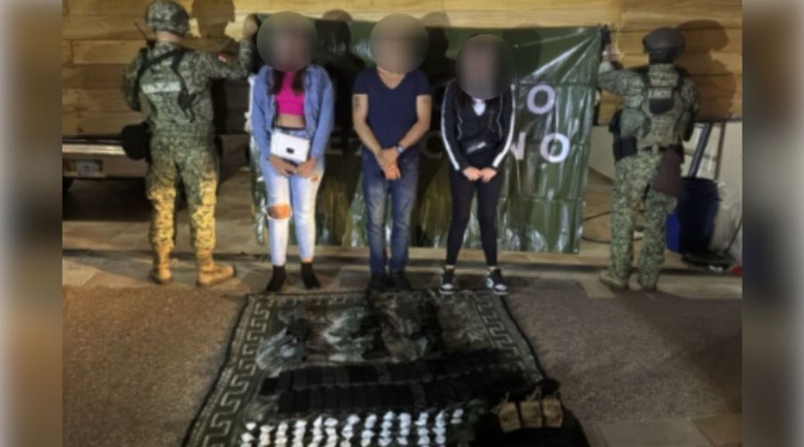 Dos elementos del ejercito mexicano sostienen una lona y presentan a 3 personas (2 mujeres y un hombre), detenidas porque se les encontraron armas, droga y material delictivo, los cuales se presentan en la parte inferior de la imagen