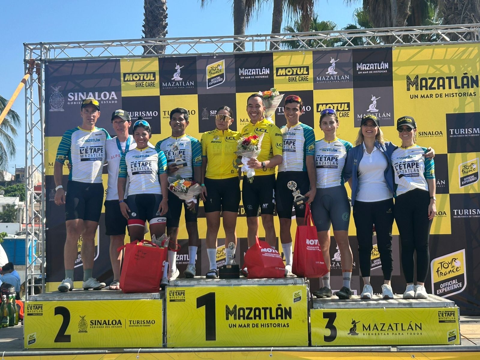 Diego Méndez y Andrea Martínez arriba del podio luego de coronarse en L’Etape Mazatlán by Tour de France, junto a otros competidores, cargando flores y premios de la competición.