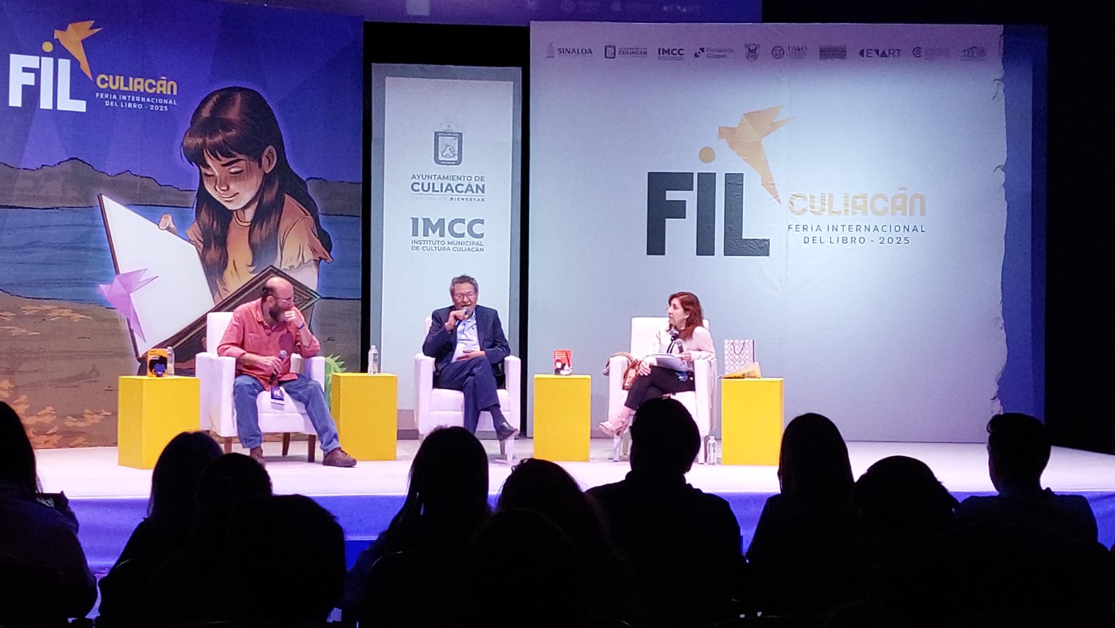 Eduardo Antonio Parra y Elizabeth Moreno y Élmer Mendoza sentados en sillones arriba del escenario de la FIL Culiacán 205, por debajo se ve al publico observando la presentación y por detrás el logotipo de la FIL y una animación de una niña leyendo un libro