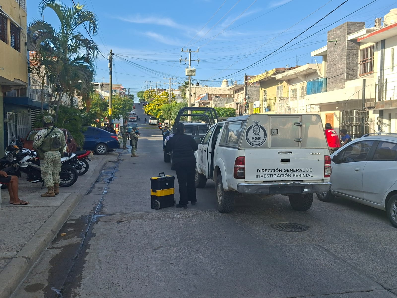 Unidad de la Fiscalía General del Estado estacionada en mitad de la calle y un elemento de la misma vestida de negro abajo de la unidad con una maleta a un lado, alrededor elementos del Ejercito Mexicano asegurando la zona del incidente