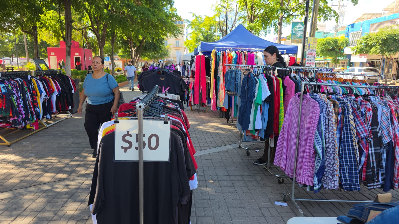 Varios puestos de ropa alrededor de una plaza, teniendo de frente los precios a los que se venden y personas transitando por la plaza.