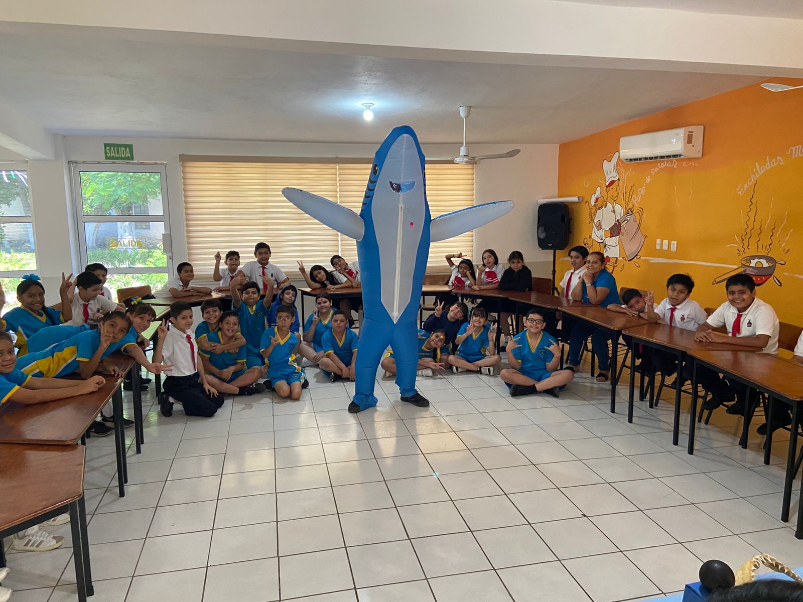 Alumnos de primaria sentados, una botarga de un delfín,