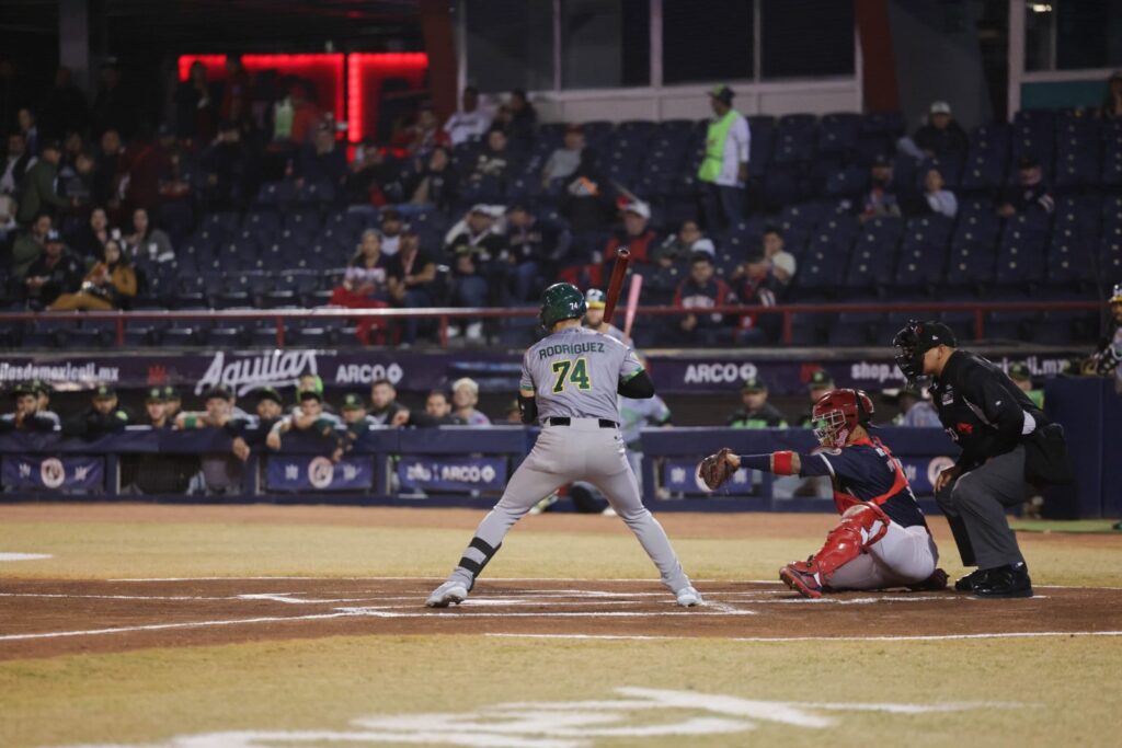 Jugador de Cañeros de Los Mochis de espaldas en la caja de bateo, en posición de impacto, por detrás el cátcher de Águilas de Mexicali esperando recibir la pelota lanzada y a su vez el ampáyer observando la dirección del lanzamiento.