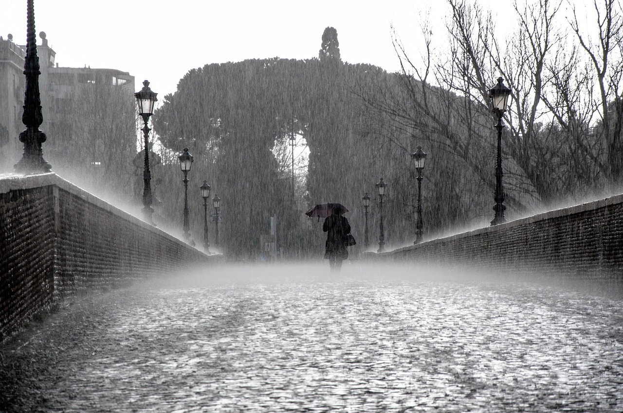 Persona caminando por la calle vestida de negro con un paraguas en la mano y con una lluvia fuertes cayendo, un cielo muy cerrado por las nubes