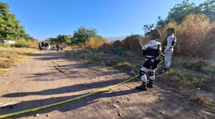 Elementos policiacos observando a personas alrededor de un cadaver cubierto con una sabana en una zona tipo baldío