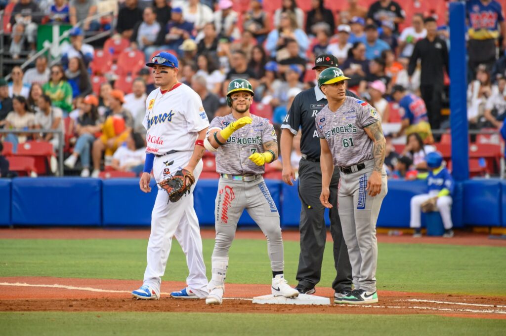 Marco Jaime (Cañeros) con los brazos hacia el frente en primera base junto a su coach que platica con el y el jugador de Charros de Jalisco que defiende la base local, por detrás el ampáyer 
