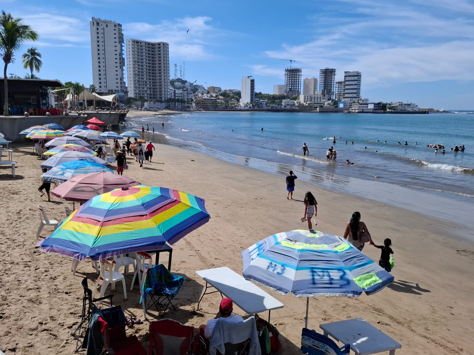 Playa de Mazatlán con buena afluencia de bañistas, se ve a algunos en la zona de la arena descansando debajo de las sombrillas y a otros bañándose en el agua