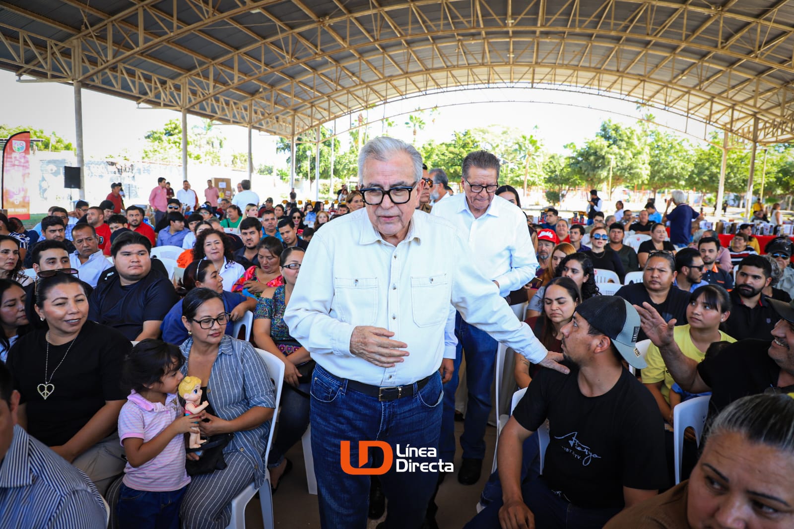 El gobernador de Sinaloa Rubén Rocha Moya caminando por en medio de una multitud sentada bajo una techumbre, mientras coloca su mano sobre el hombro de un ciudadano de manera amistosa