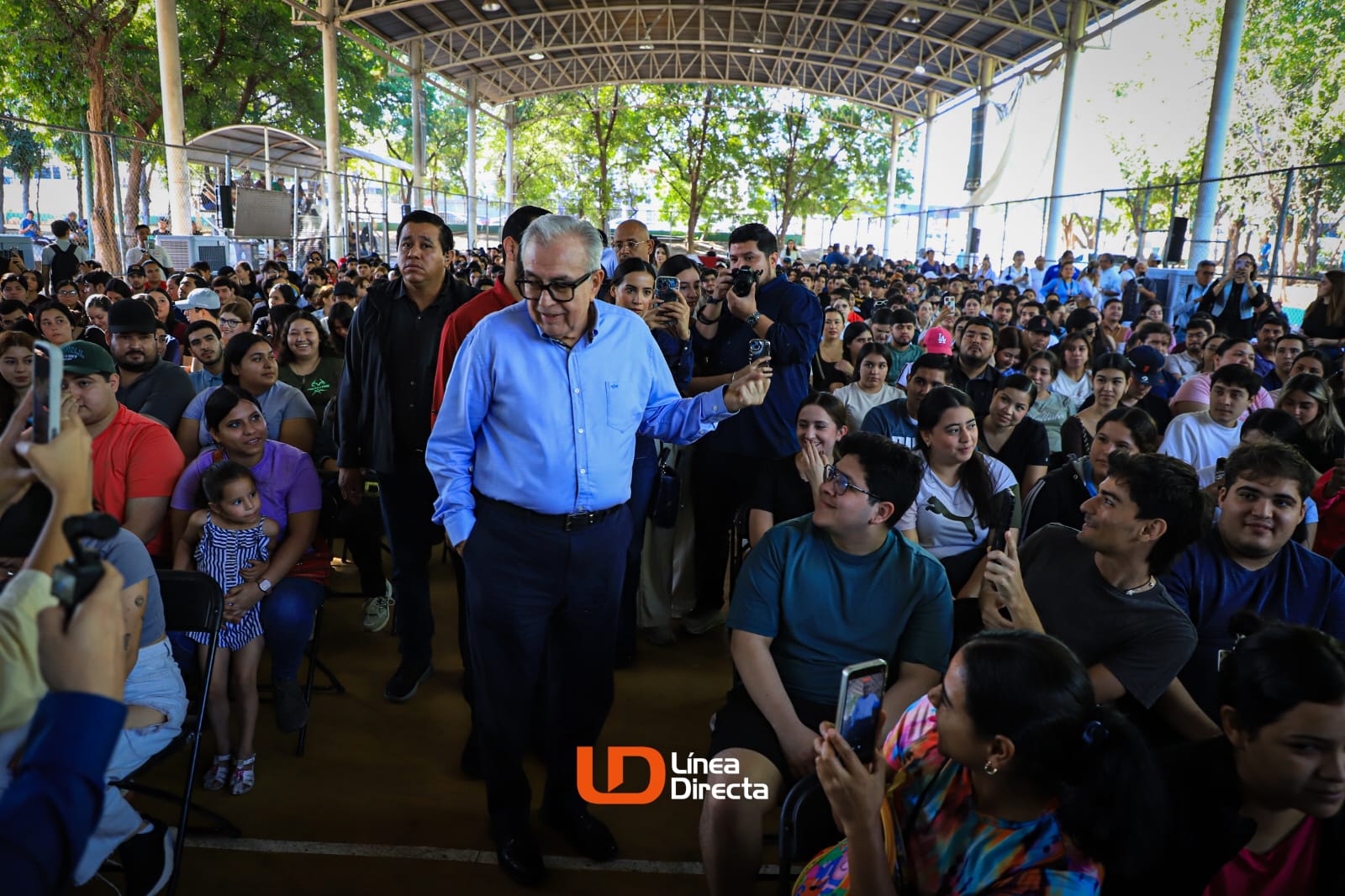 Gobernador Rocha caminando y saludando a jóvenes sentados que lo miran y lo graban con sus celulares.