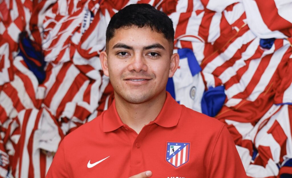 Jugador mexicano Ronaldo Nájera dentro de las oficinas del Atlético de Madrid, portando el uniforme del club español y señalando al escudo 