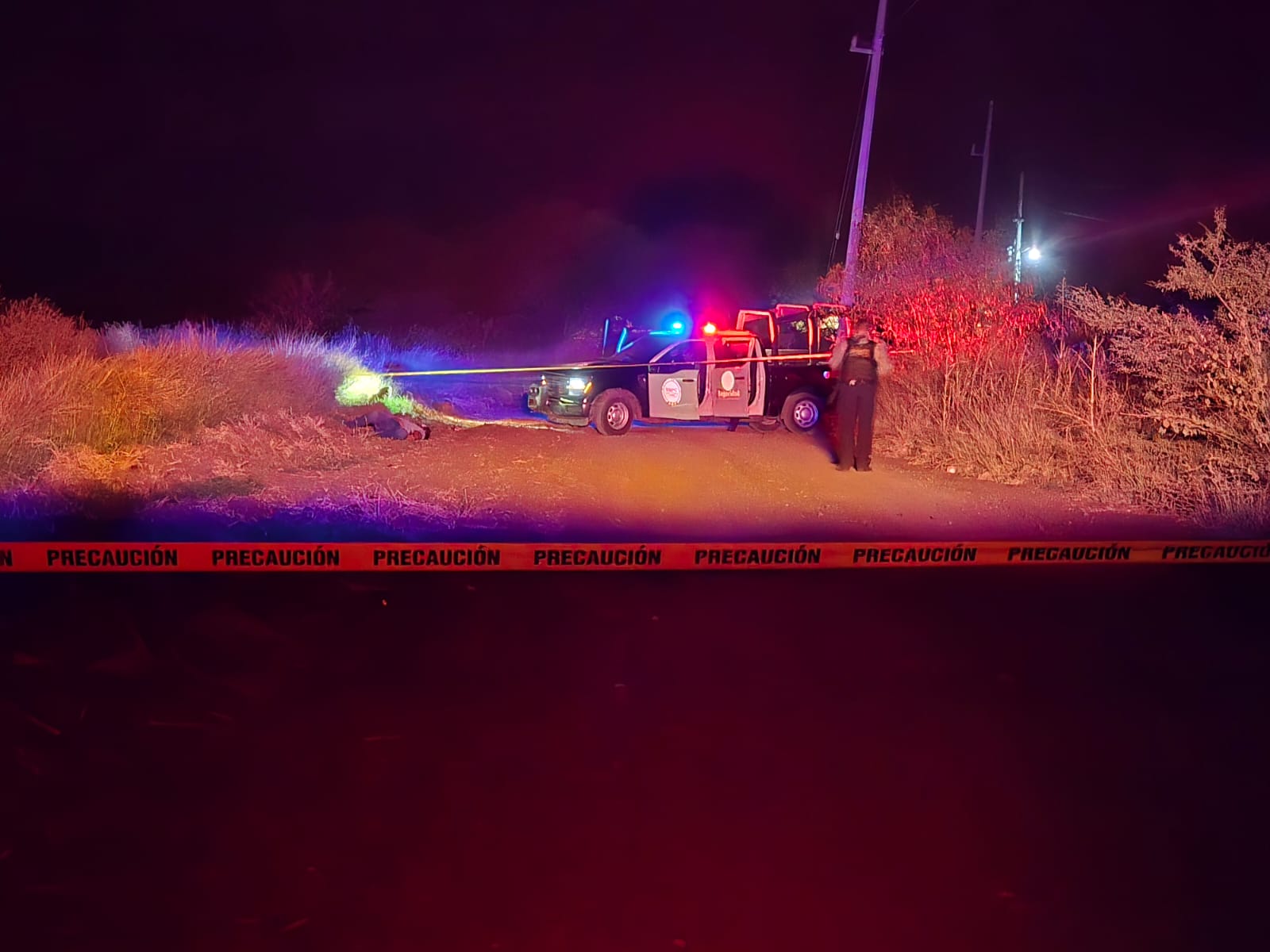 Escena nocturna de un operativo policiaco en camino rural acordonado con cinta de precaución, donde agentes resguardan el área tras el hallazgo de una persona tendida entre la maleza