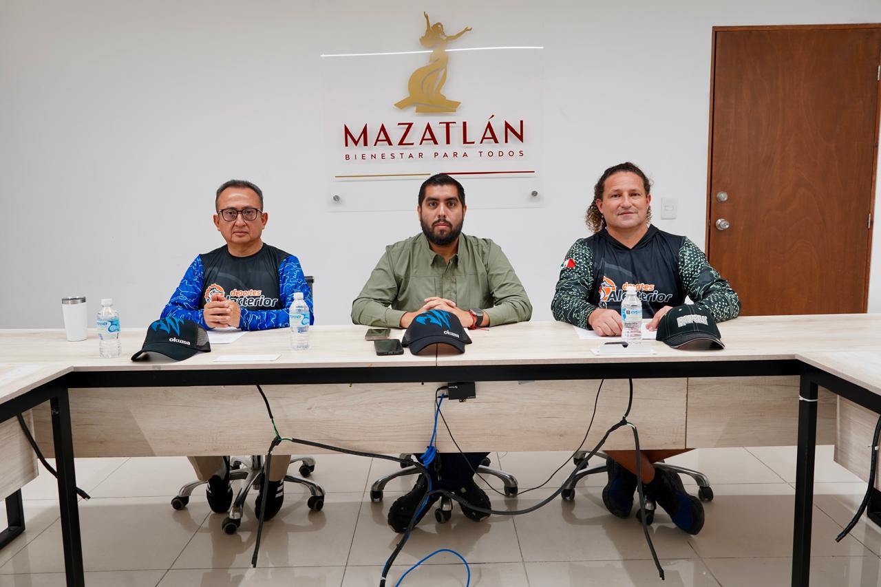 Tres autoridades de la competencia anunciada entre ellas coordinador de Turismo del Gobierno Municipal, sentados en una mesa dando declaraciones a los medios de comunicación, por detrás de ellos el logo del gobierno de Mazatlán