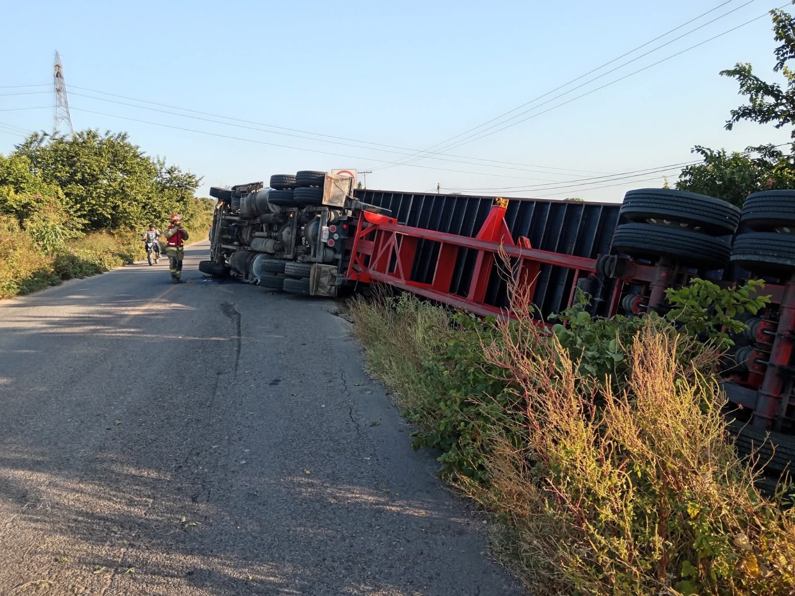 Un tráiler volcado sobre la carretera, mientras que un bombero acude a la ayuda del accidentado, a su vez un transeúnte en bicicleta va pasando por el lugar