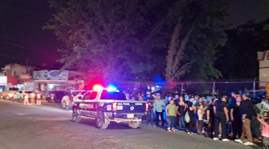 Una unidad de Policía con las torretas encendidas estacionada en una calle donde hay un gran grupo de personas alrededor.