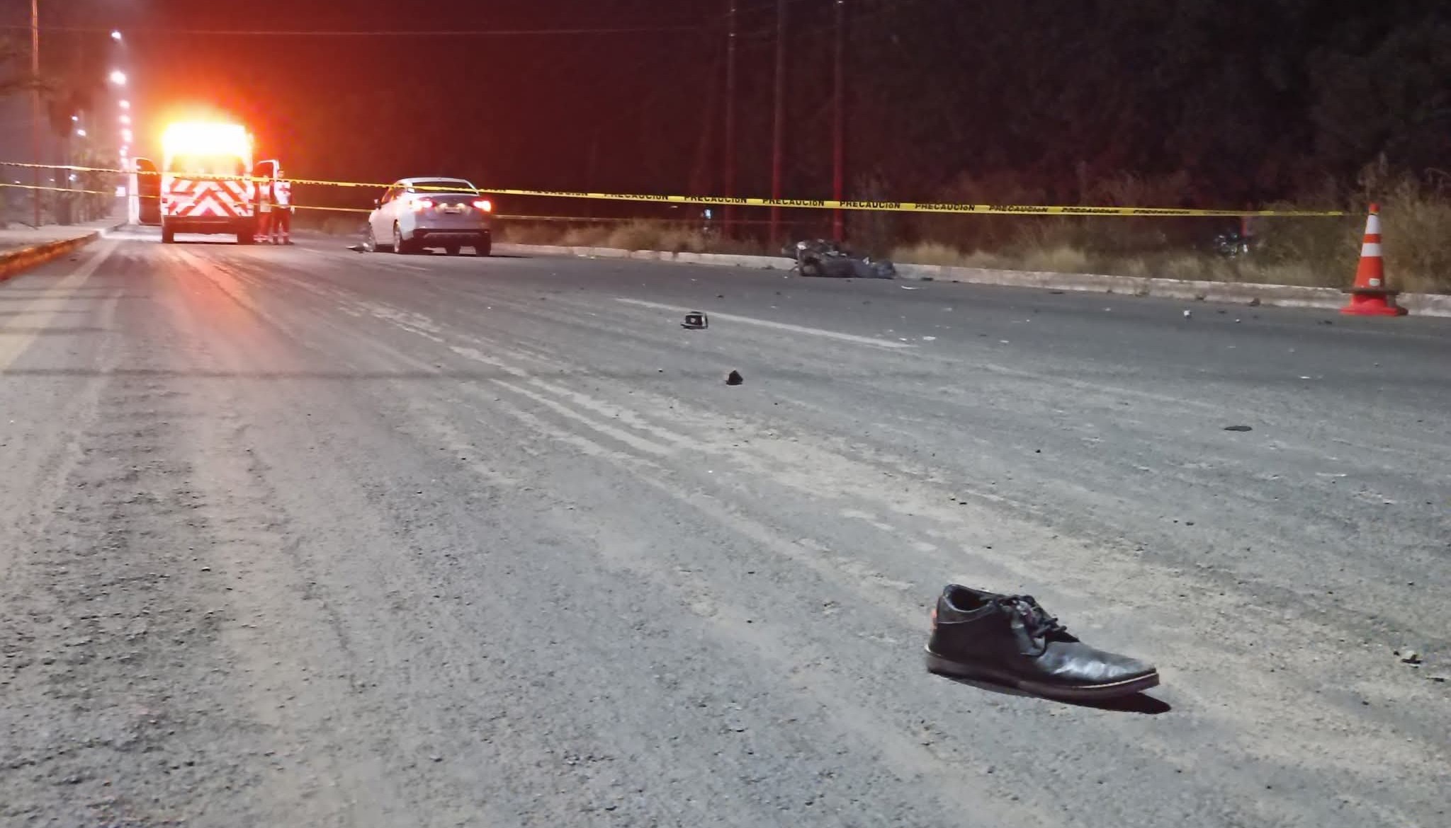 Zapato negro tirado en la calle, al fondo una cinta amarilla de precaución junto a un vehiculo color gris y una unidad de la Cruz Roja