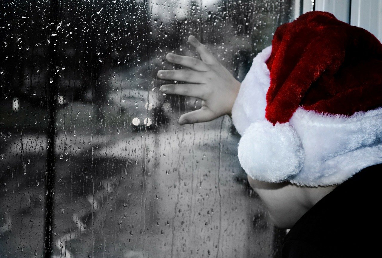 Joven con gorro navideño viendo por ventana durante noche lluviosa