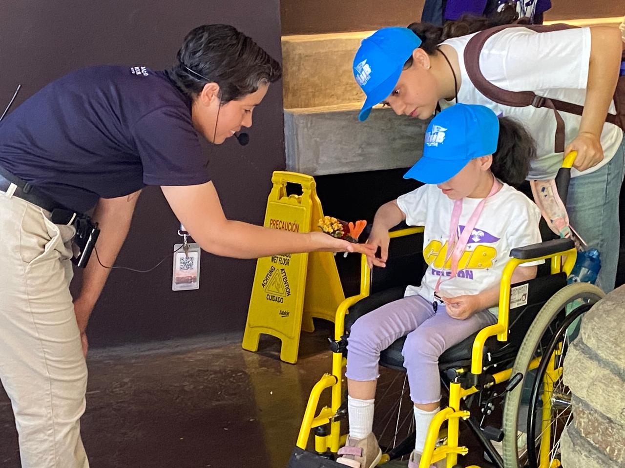 Personal de Gran Acuario Mazatlán enseña objeto a niña en sillas de ruedas que sostiene una medalla
