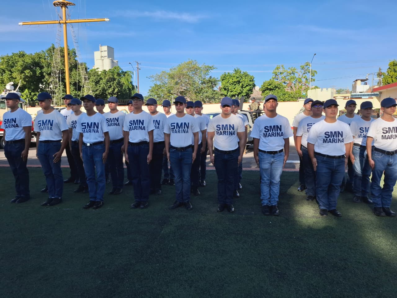 Jovenes portando una playera blanca con las letras SMN en color negro con gorra, pantalones oscuros y zapatos negros durante un acto de graduación, cielo azul
