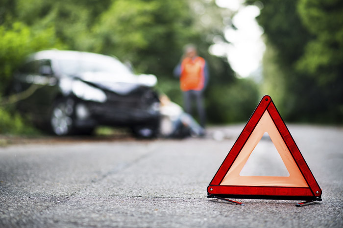 Signo de precaución en forma triangular en colores rojo y naranja, al fondo se observa un accidente de un carro en una calle