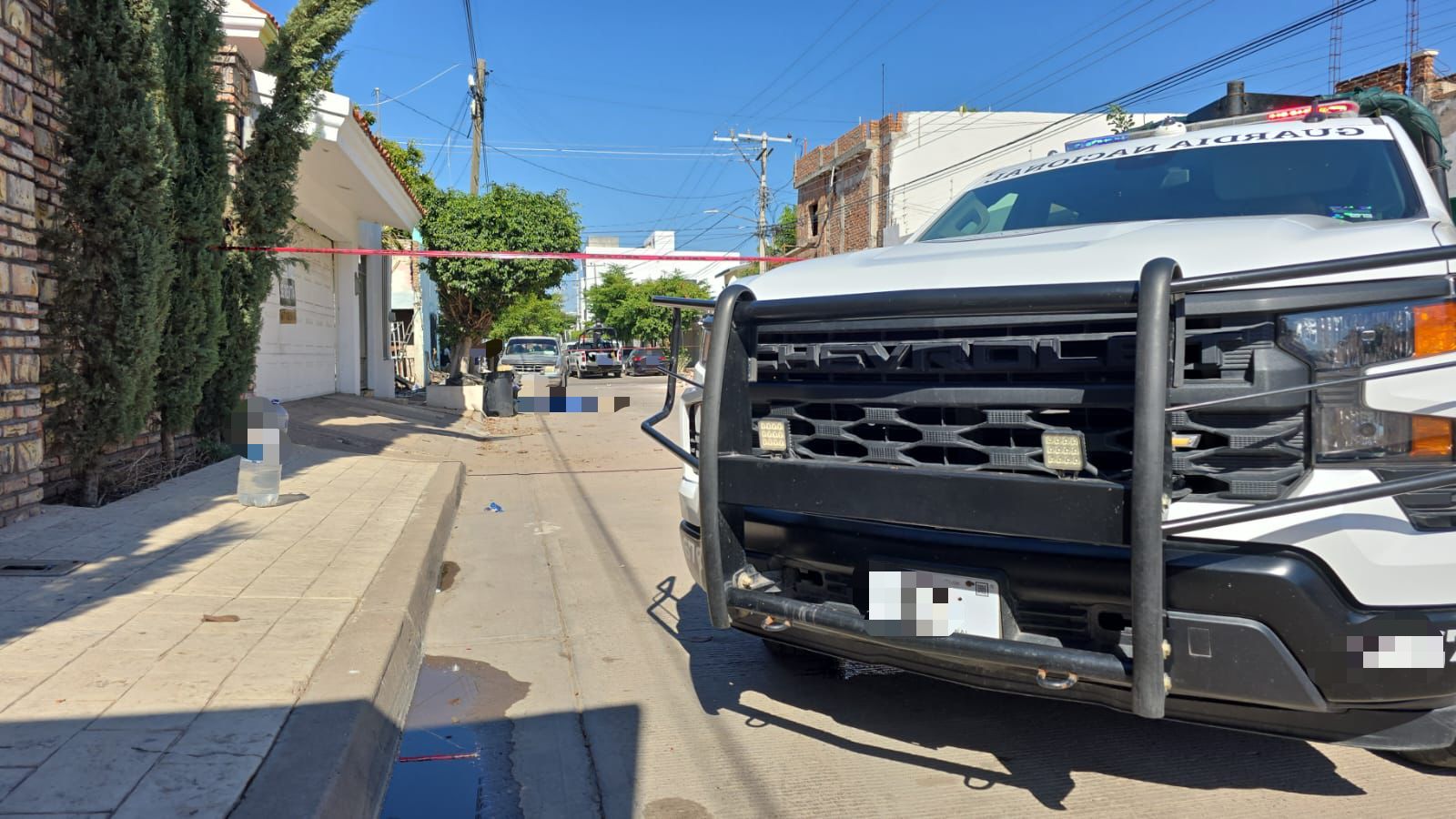 Unidad policiaca en una calle, al fondo se observa una cinta de precaución y un cadaver tirado con una sábana azul sobre puesta