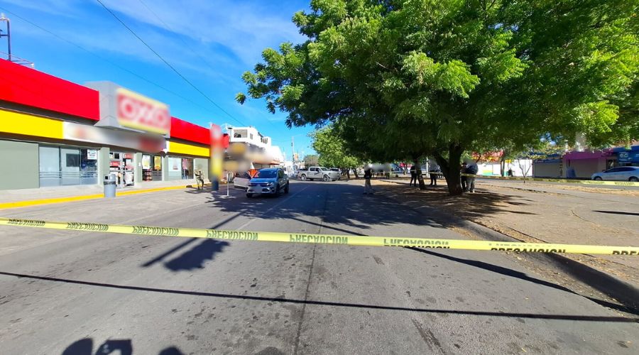 Carro baleado en medio de una calle frente a una tienda de autoservicio en color rojo y amarilllo
