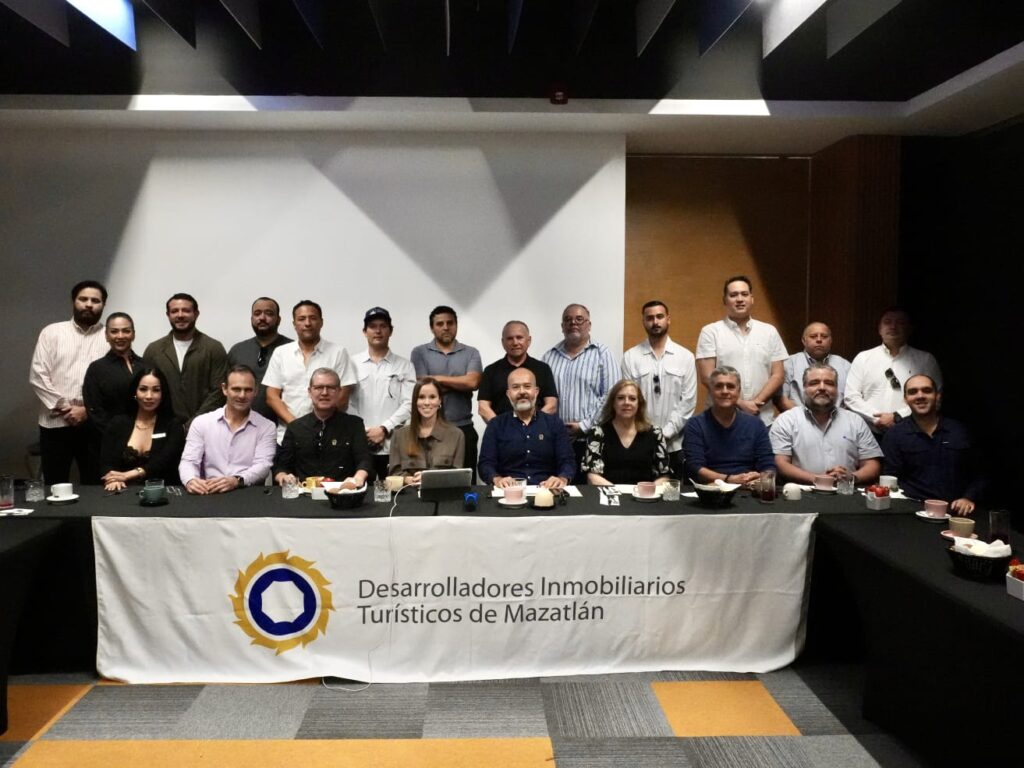 Personas de pie y sentadas en un foro con una mesa que tiene una manta con la leyenda de Asociación de Desarrolladores Inmobiliarios Turísticos