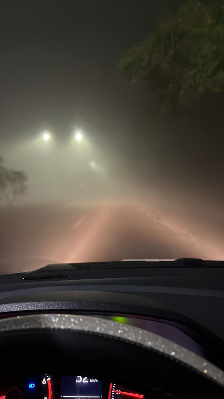 Tablero de un carro con las luces altas iluminando el camino cubierto por bancos de niebla, al fondo se observan luminarias entre la niebla