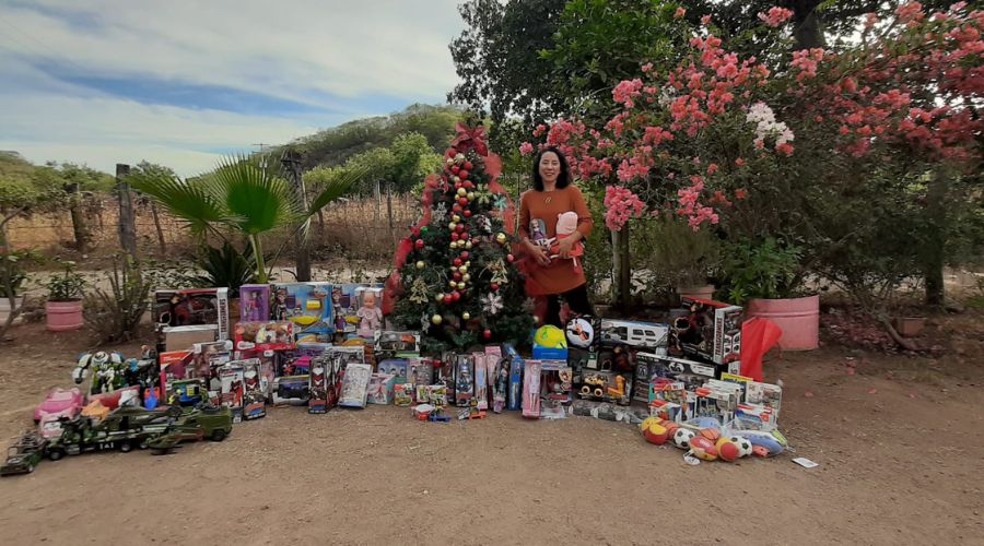 Mujer de pelo corto, blusa café y pantalon negro en un lugar tipo campo con regalos en sus brazos y en el suelo