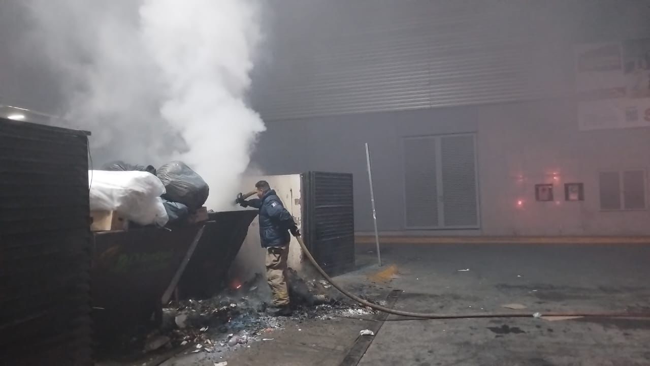 Persona con una manguera cerca de un contenedor de basura con bolsas saliendo humo