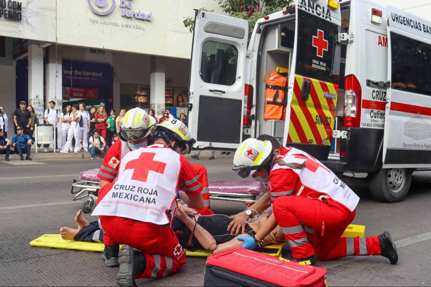 Personal de cuerpo de emergencia auxiliando a una persona que está tirada en la calle, atrás una unidad de la Cruz Roja