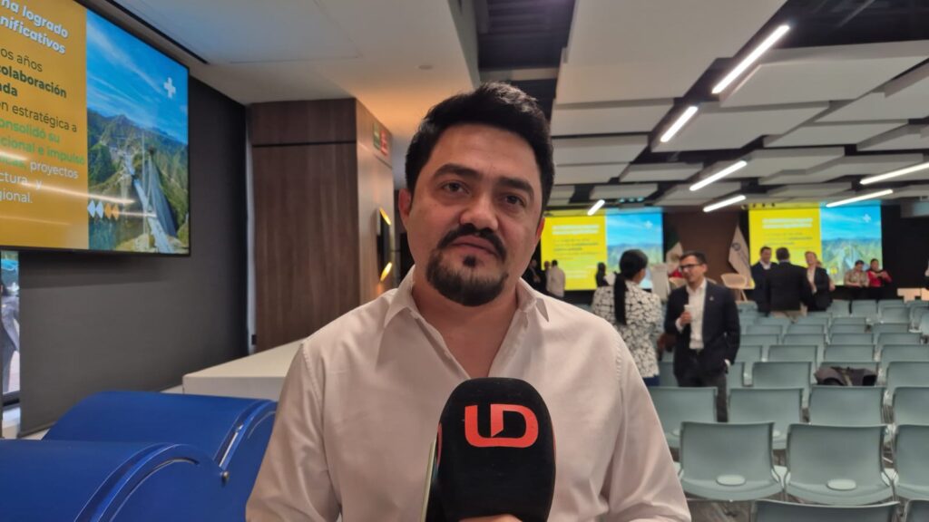 Hombre de cabello negro y camisa color claro viendo hacia el frente y un micrófono negro con las letras LD en color naranja, en un foro/salón