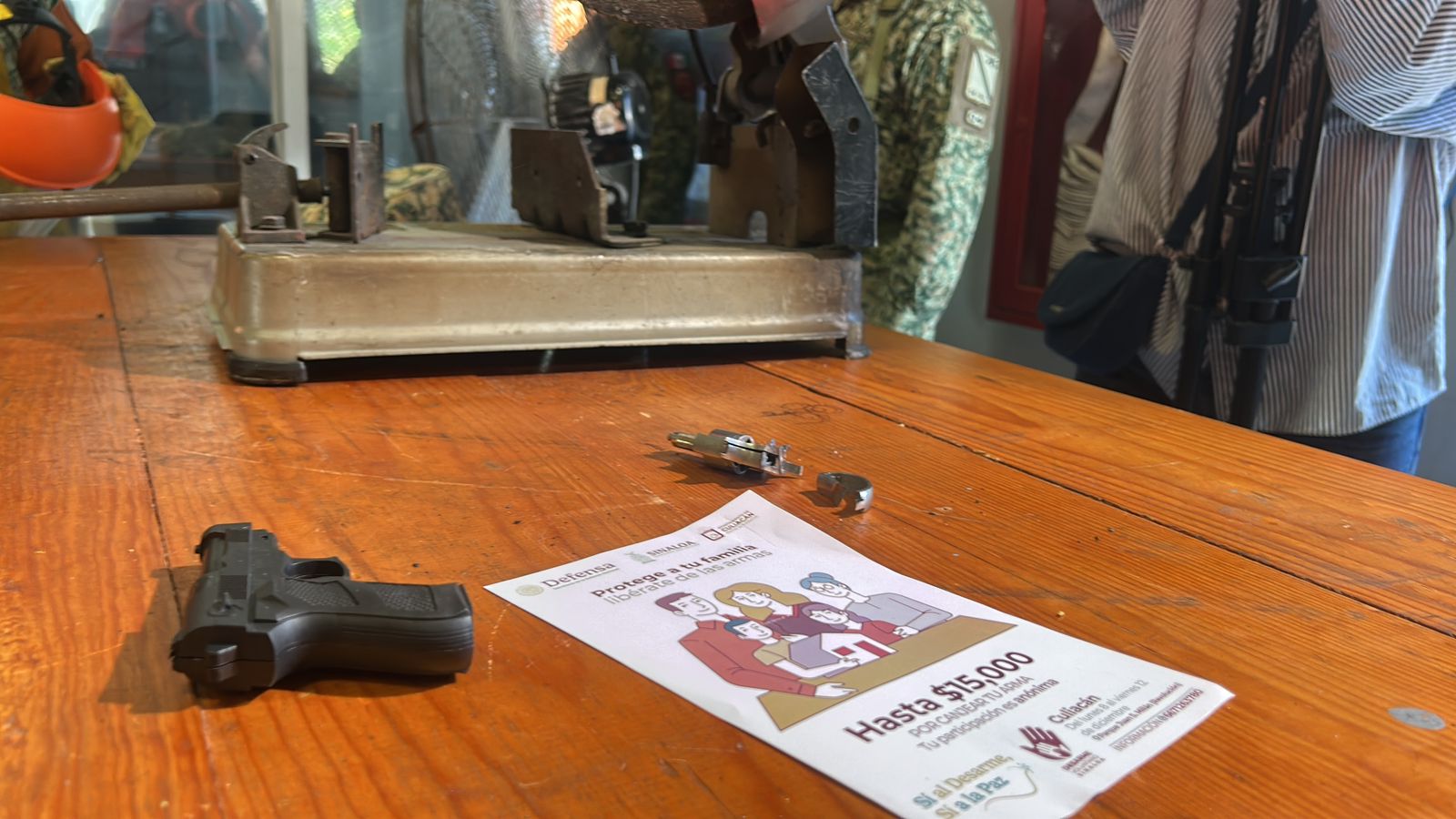 Arma de fuego sobre una mesa junto a un volante de la campaña Desarme Voluntario Sinaloa 2025