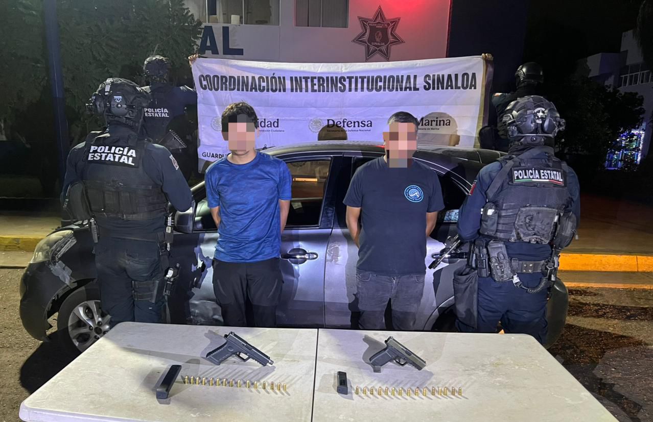 Elementos de la policía estatal sosteniendo una lona junto a 2 civiles detenidos con las manos en las espaldas, en una mesa blanca hay armas que fueron aseguradas