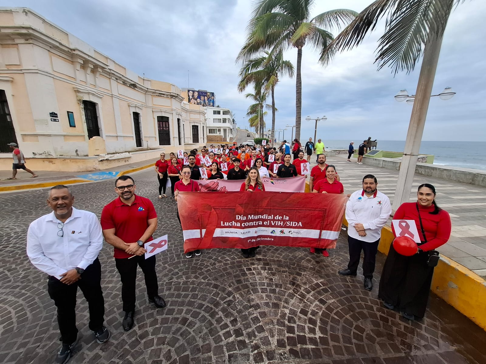 Personas portando ropa color rojo y blanco en una marcha en una calle con cielo nublado y palmeras