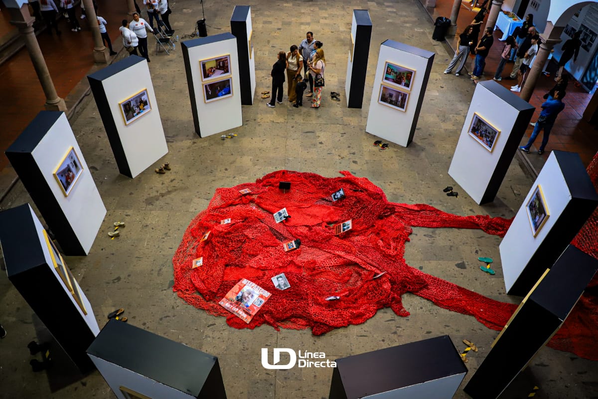 Murales colocados en circulo donde están colocadas fotos de personas desaparecidas, en medio una tela roja donde están mas fotografías de personas desaparecidas y alrededor personas mirando la exposición