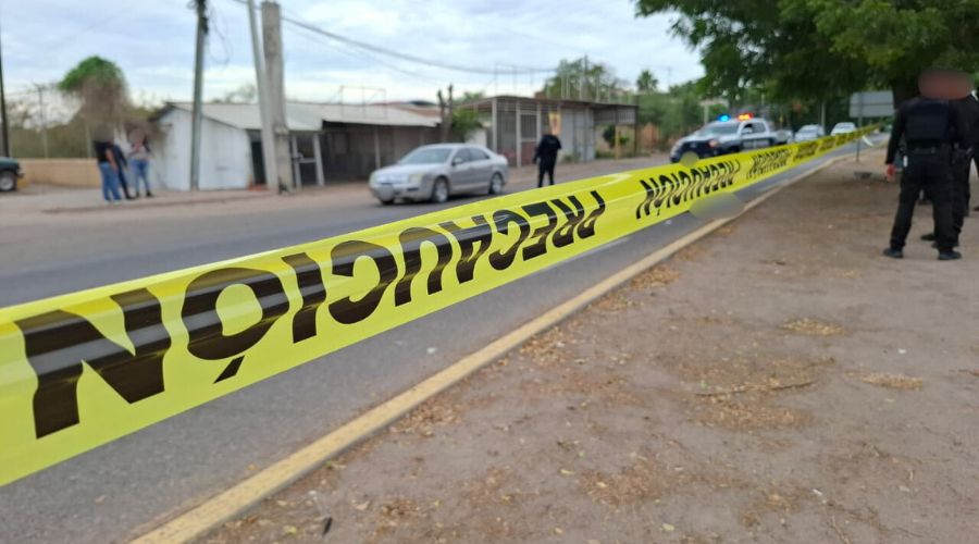Cinta amarilla de precaución en una calle donde hay un cadáver tirado en la calle, con policías y una unidad policiaca