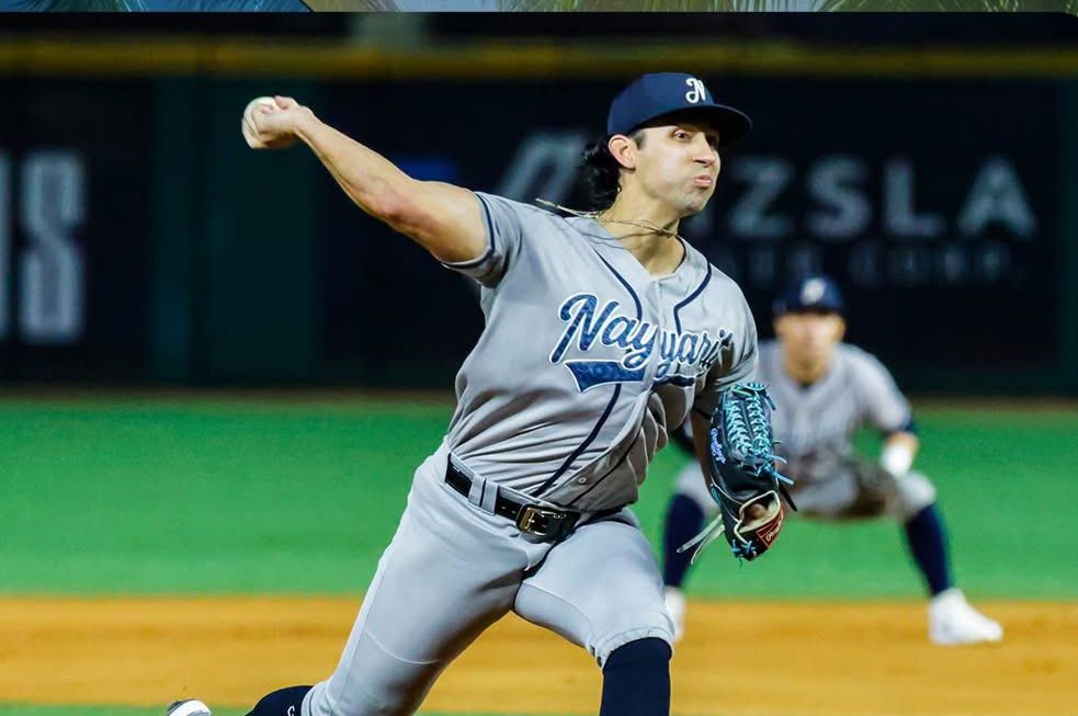 Jugador de beisbol con uniforme color gris lanzando una pelota en un estadio, al fondo se ve a otro jugador de beisbol en posición de receptor