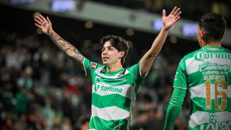Jugador de futbol en un estadio con uniforme en colores verde y blanco levantando sus brazos