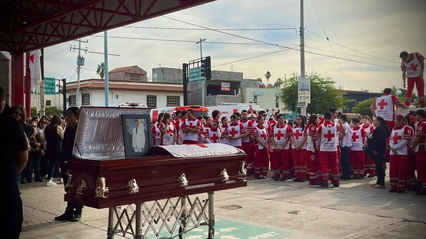Féretro con una fotografía en marco en primer plano. Al fondo grupo de personas paramédicos de Cruz Roja en Guasave