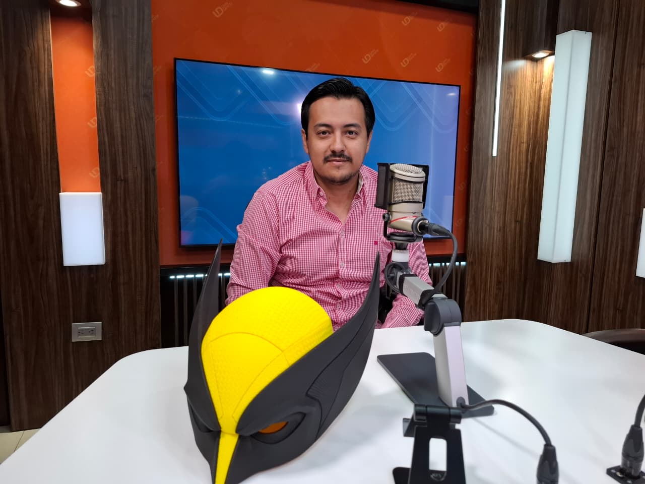 Joven en estudio de noticiero de radio frente a un micrófono y una máscara