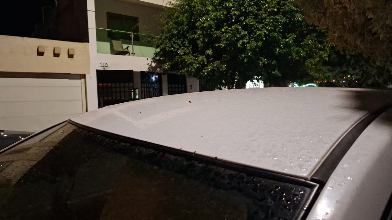 Gotas de agua arriba de un carro blanco y al fondo se ven casas y un arbol