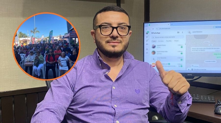 Hombre con camisa morada y lentes sentado viendo al frente levantando el dedo pulgar, atrás hay una computadora y en un círculo personas sentadas en una reunión