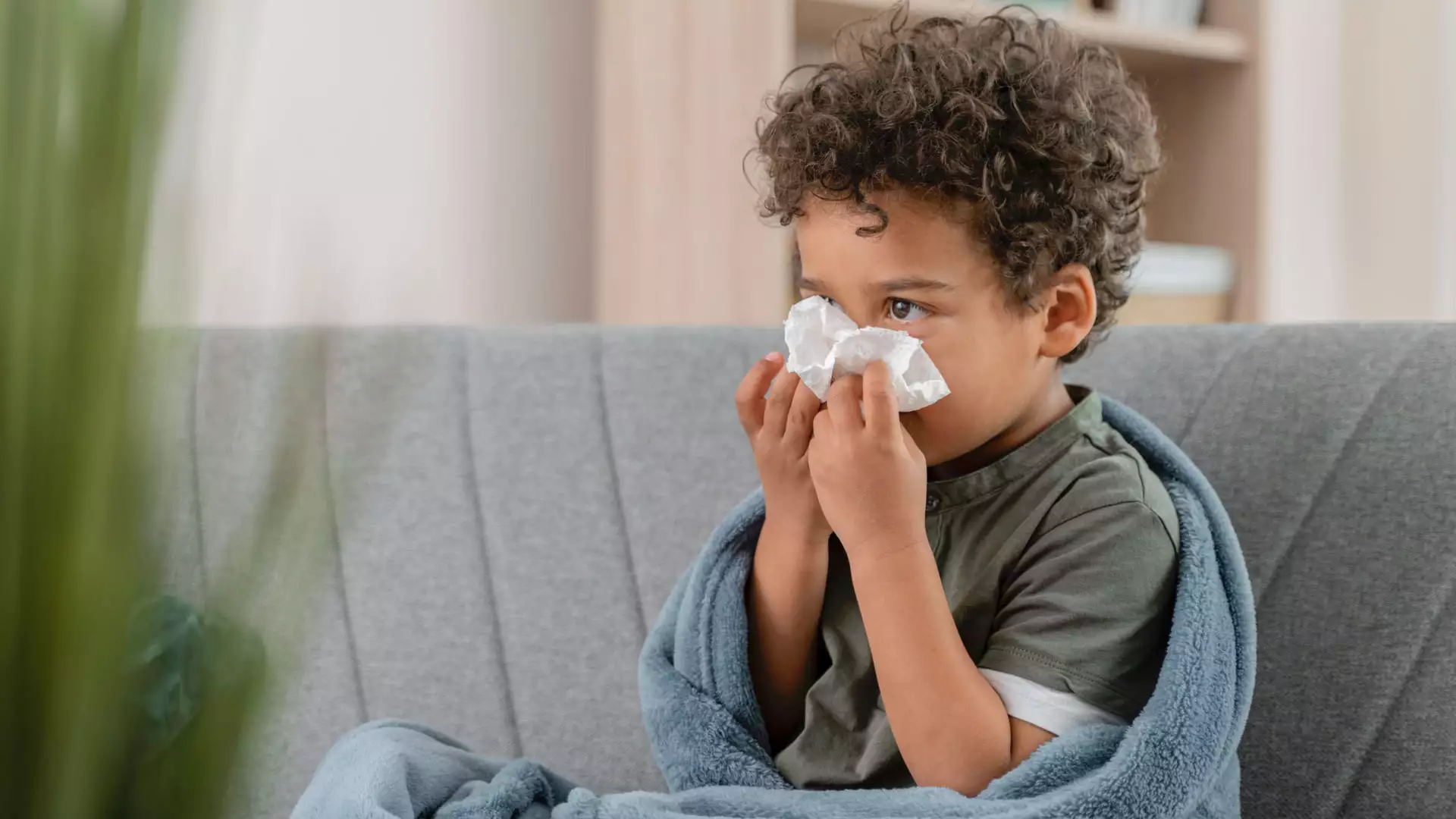Un niño pequeño sentado en un sillón con una cobija y sonándose la nariz