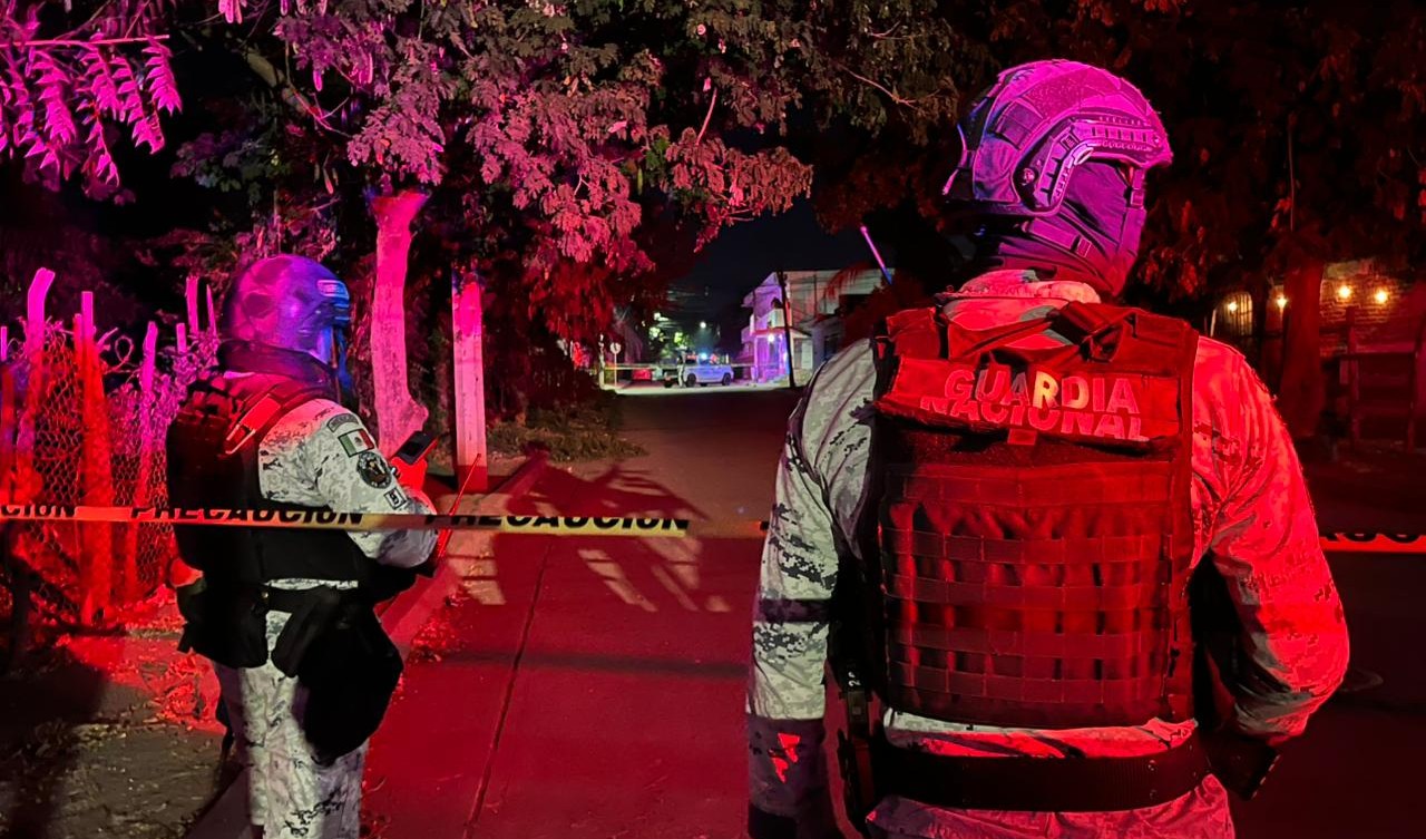 Dos elementos de la Guardia NAcional de espaldas en una calle con una cinta amarilla de precaución, al fondo se observa una unidad de la Guardia Nacional