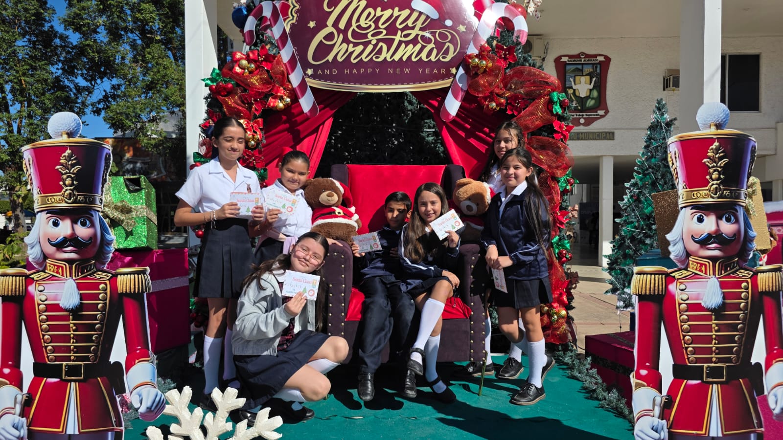 Niños con uniformes de escuela en una tarima sentados en medio de regalos y adornos navideños con cartas en sus manos