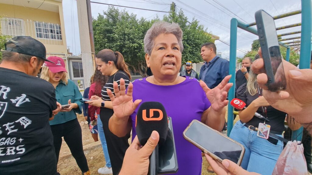 Persona adulta de pelo corto con blusa morada hablando a muchos micrófonos