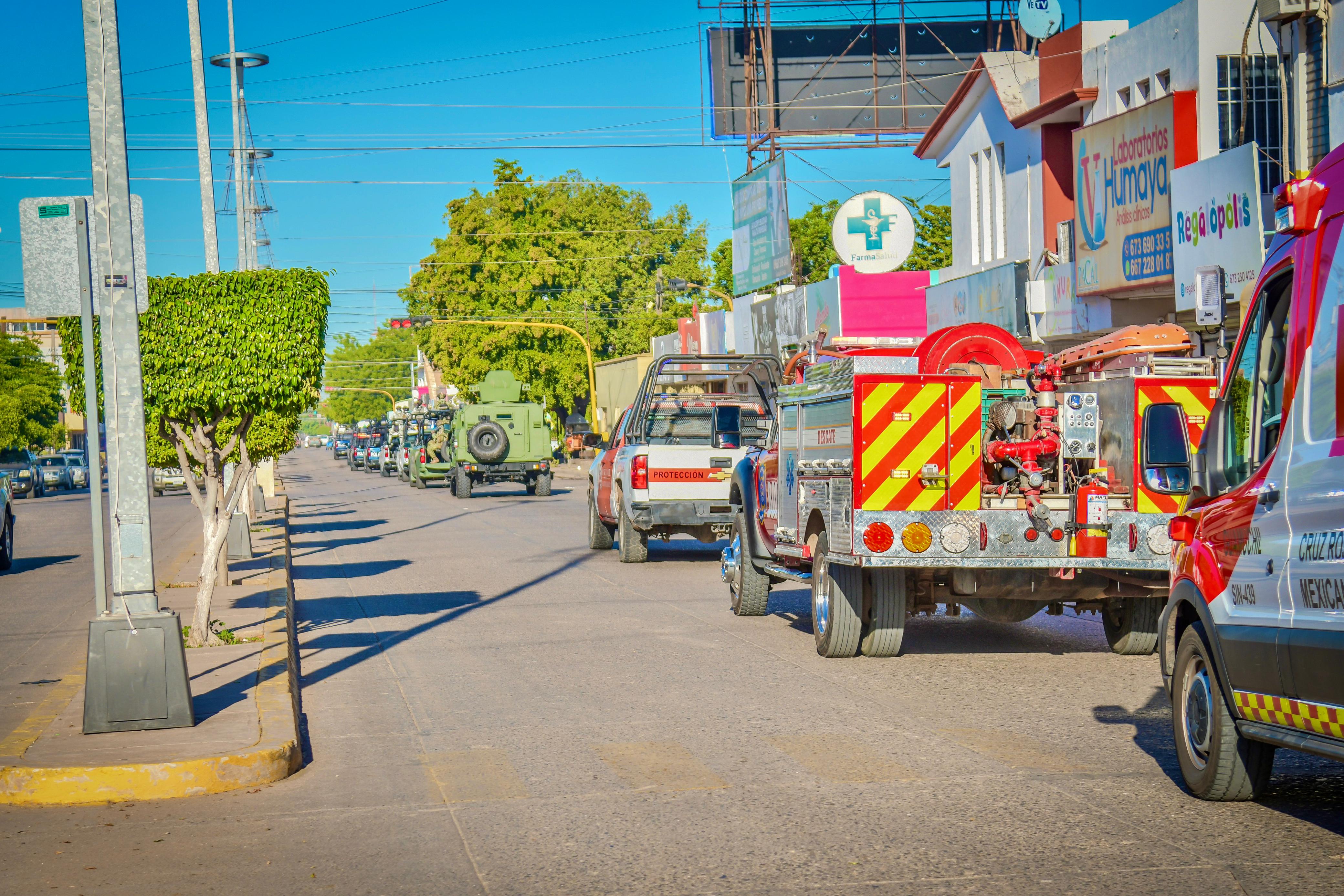 Unidades de diferentes corporaciones como bomberos, policìa, cruz roja, en una calle con arboles y cielo azul